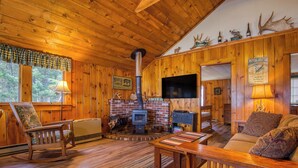 A cozy cabin interior with wooden walls, a fireplace, a rocking chair, a sofa, and a television. - 🐾 Mooselodge | Cozy Cabin on Rangeley Lake, Pet Friendly (Rangeley)