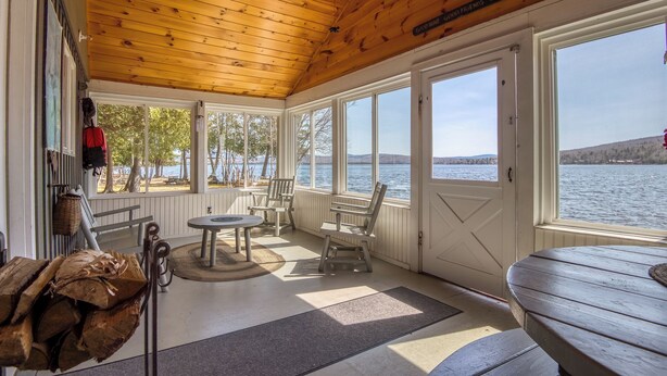 A covered porch with wooden ceiling, white walls, and large windows overlooking a lake. There are chairs, a table, and a bench. -   🐾 Mooselodge | Cozy Cabin on Rangeley Lake, Pet Friendly (Rangeley)