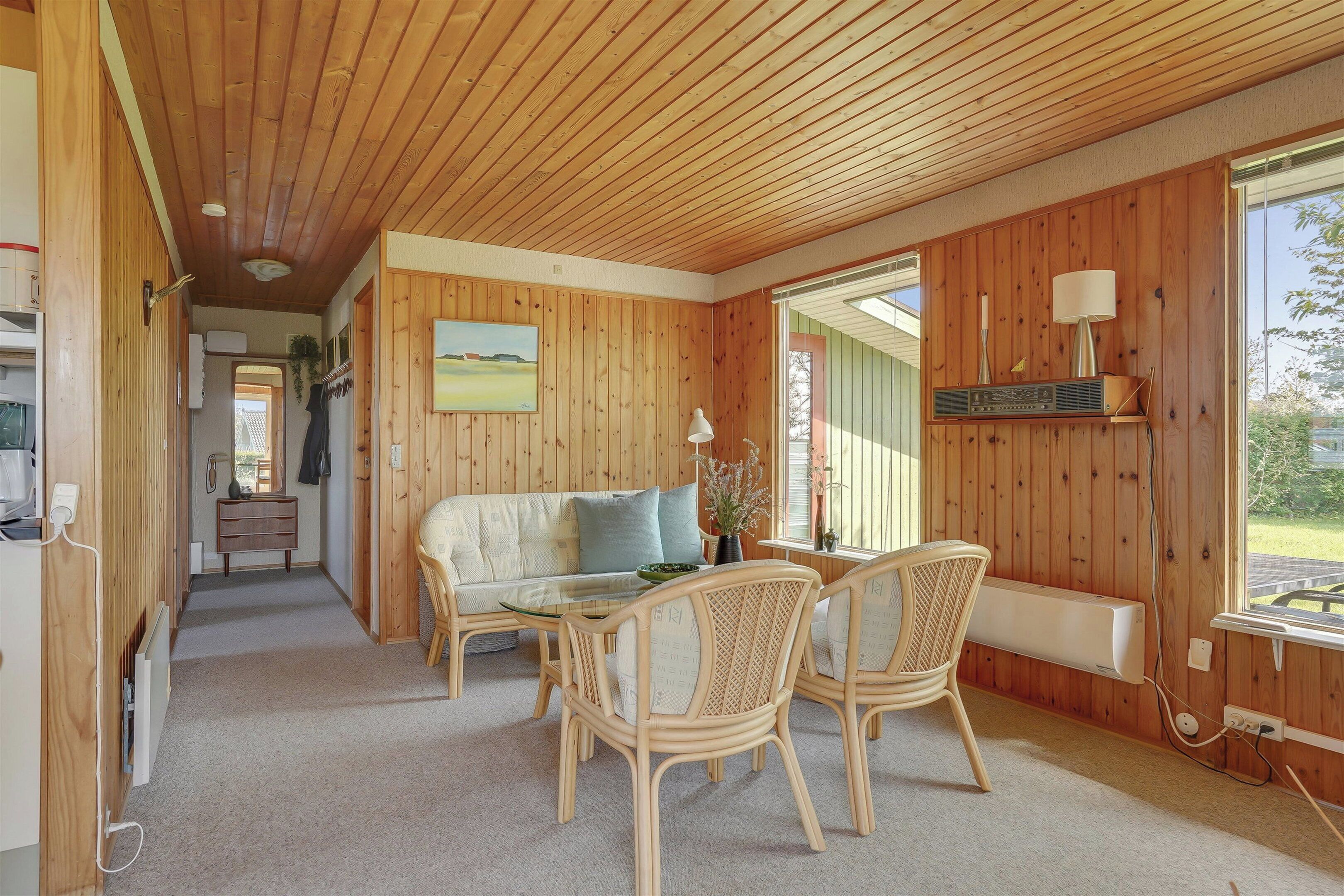 A room with wooden walls, a sofa, a dining table with chairs, a radiator, and a window with a view.