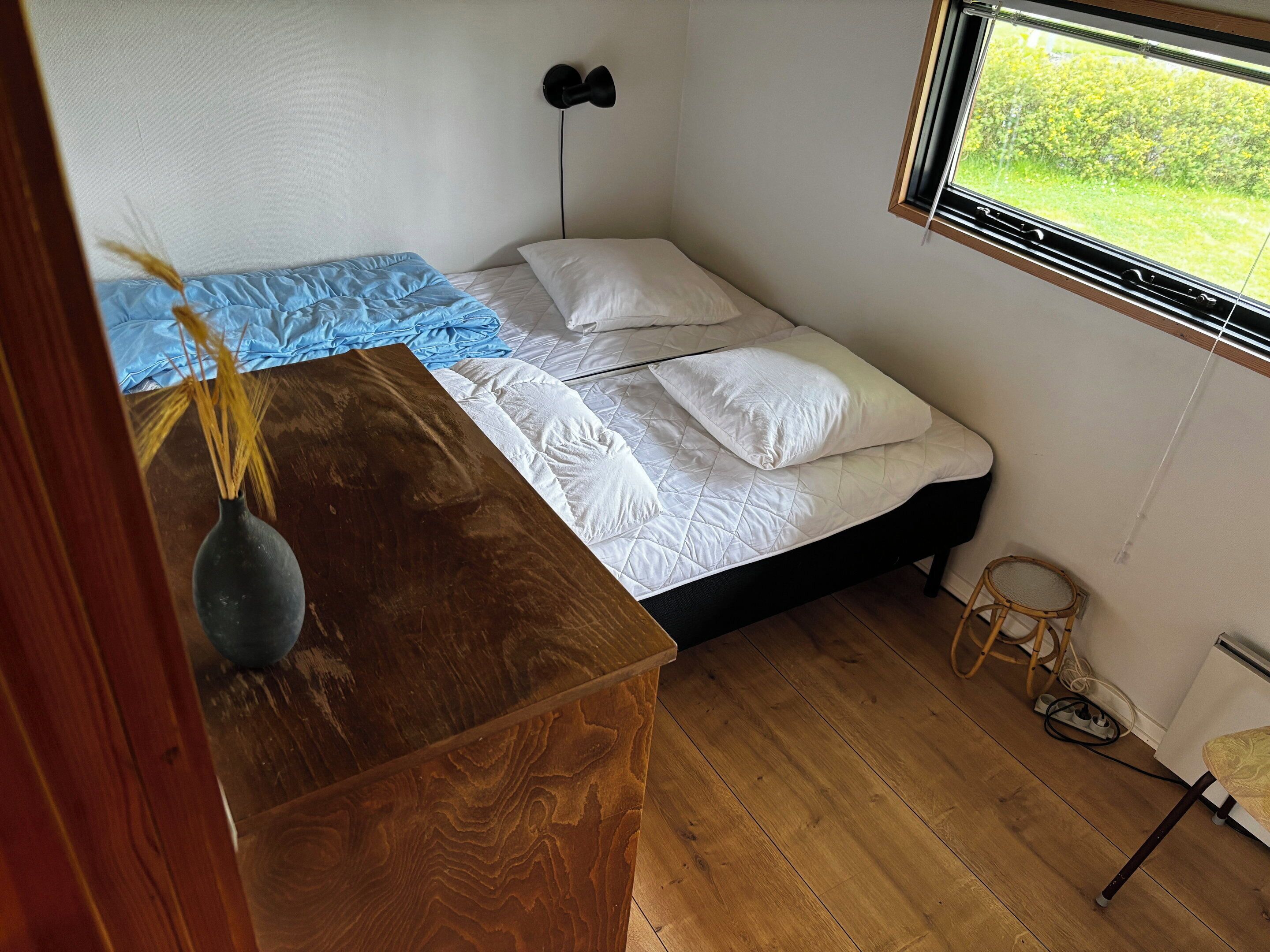A small bedroom with a bed, a bedside table, a window, and a view of greenery outside.