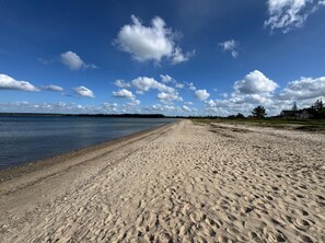 Une plage de sable fin, une étendue d’eau calme, un ciel dégagé et des maisons qui se dessinent à l’horizon.