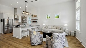 A modern kitchen with white cabinetry, a stainless steel refrigerator, and a central island. There is a seating area with a sofa and two patterned ottomans.