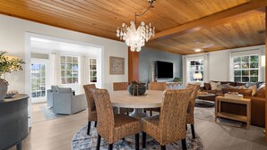 A spacious living room with a round wooden dining table surrounded by wicker chairs, a large chandelier, and a television mounted on the wall. - ⚓Dolphin Cabin! Screened Pool, Dock & More! ⚓ (Bradenton)