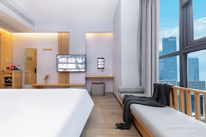 Une chambre d’hôtel avec un grand lit, un bureau avec un ordinateur, un coin salon offrant une vue sur la ville et une petite poubelle.