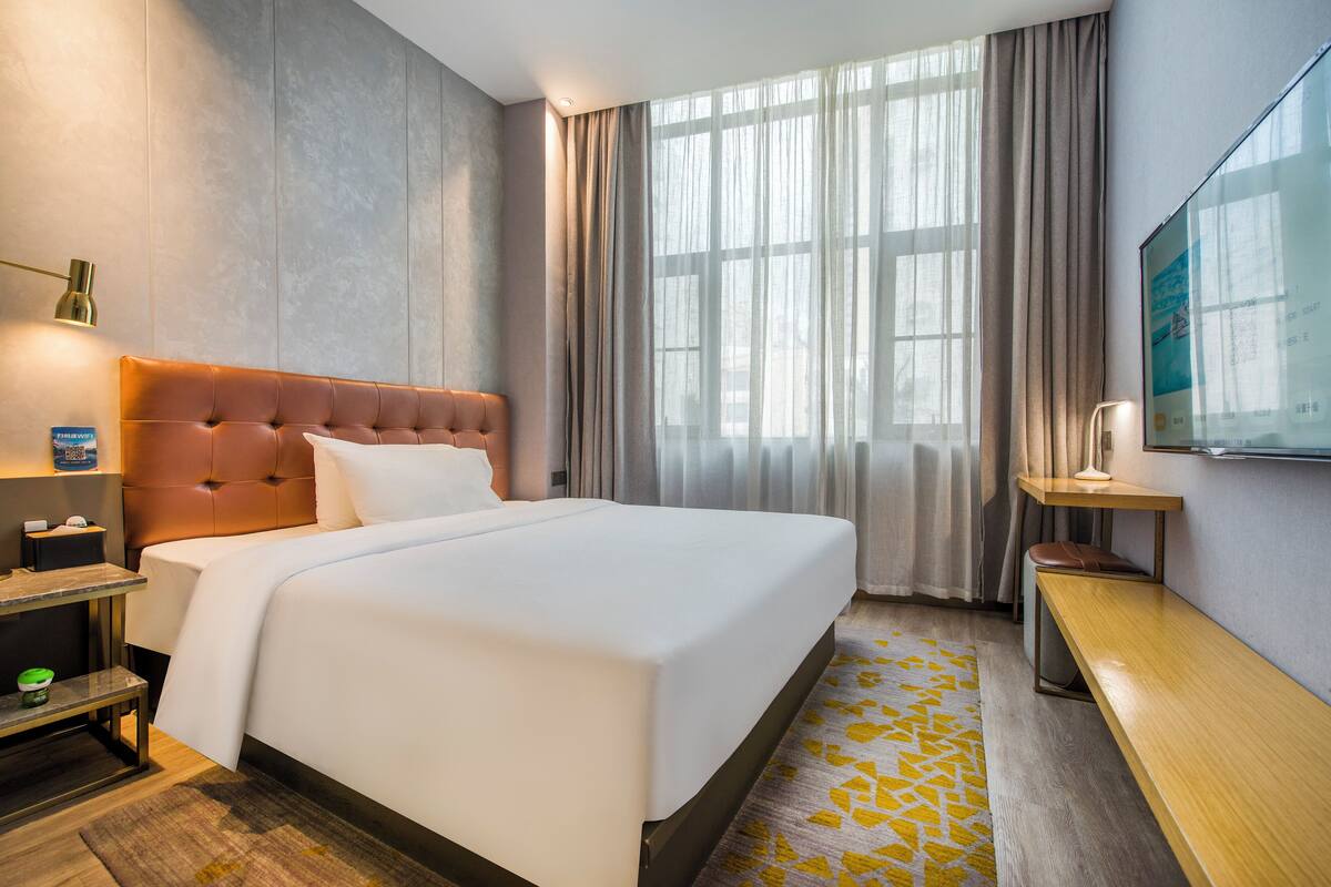 A hotel room with a large bed, a bedside table, a bench, a TV, and a window with curtains.