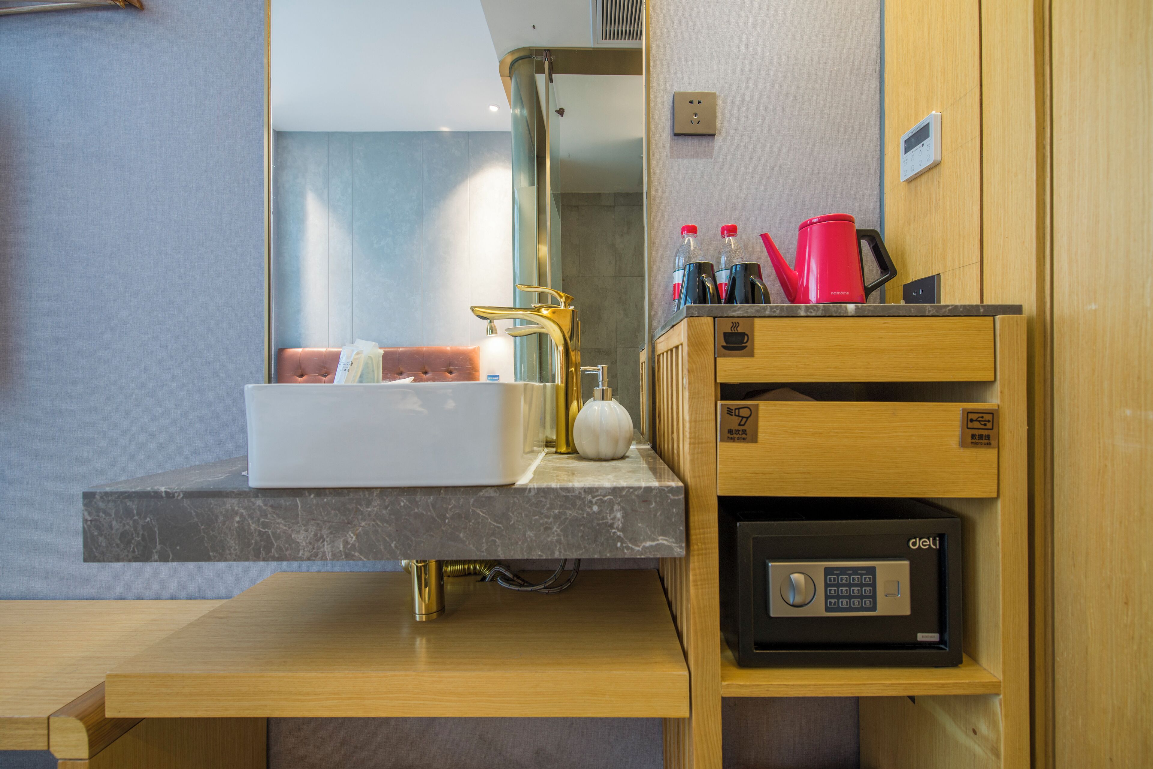 A modern bathroom with a marble sink, a gold faucet, a mirror, and a wooden vanity with a coffee maker and toiletries.