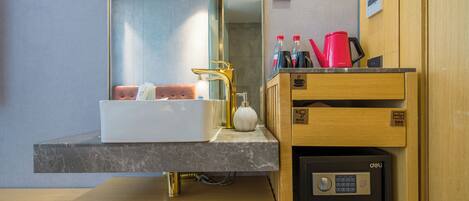A modern bathroom with a marble sink, a gold faucet, a mirror, and a wooden vanity with a coffee maker and toiletries.