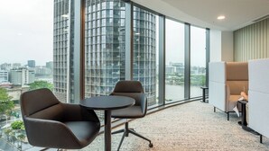 Un espacio de oficina moderno con ventanas amplias, dos sillas, una mesita y vistas a un paisaje urbano.