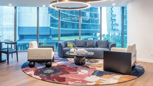 Una sala de estar moderna con un sofá gris, dos sillones, una mesa de centro y un amplio ventanal con vistas a un edificio y zonas verdes.