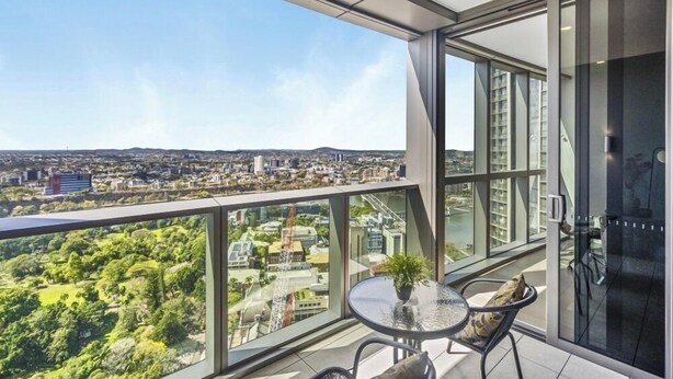 Un balcón moderno con barandilla de vidrio, una pequeña mesa con una planta y vistas a un paisaje urbano y zonas verdes.