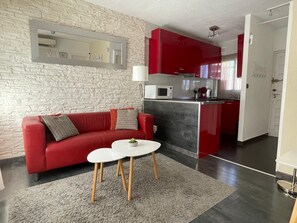 A modern living room with a red sofa, two white tables, a grey rug, a stone-textured wall, and a kitchen with red cabinets and a white microwave.