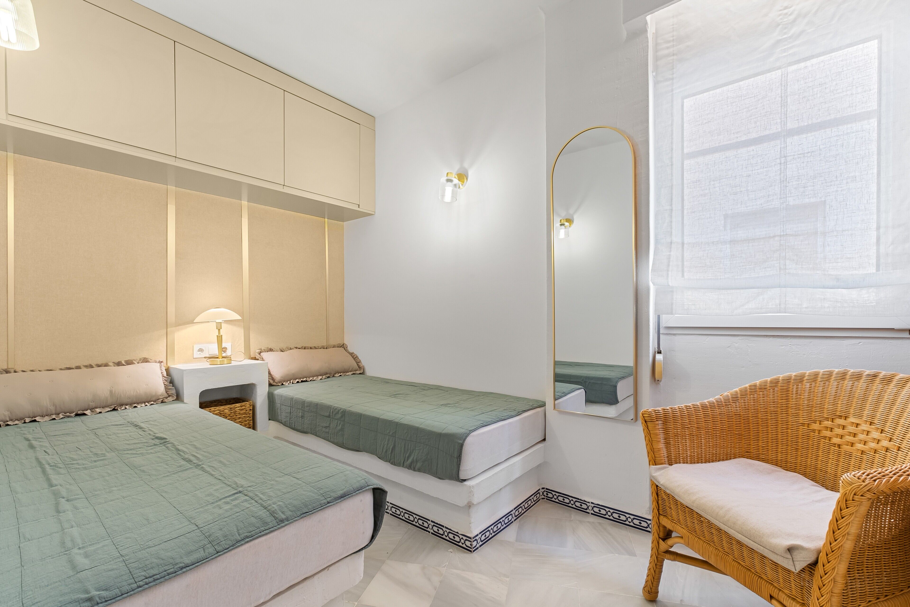 A bedroom with two beds, a wicker chair, a mirror, and a window.