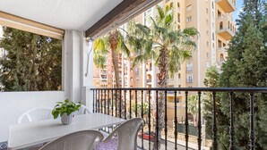 A balcony with a white table and chairs, a potted plant, and a view of a multi-story building with balconies and palm trees.