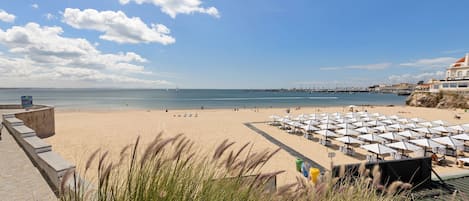 Une plage avec des parasols et des chaises longues, un ciel dégagé et une jetée qui se profile à l’horizon.