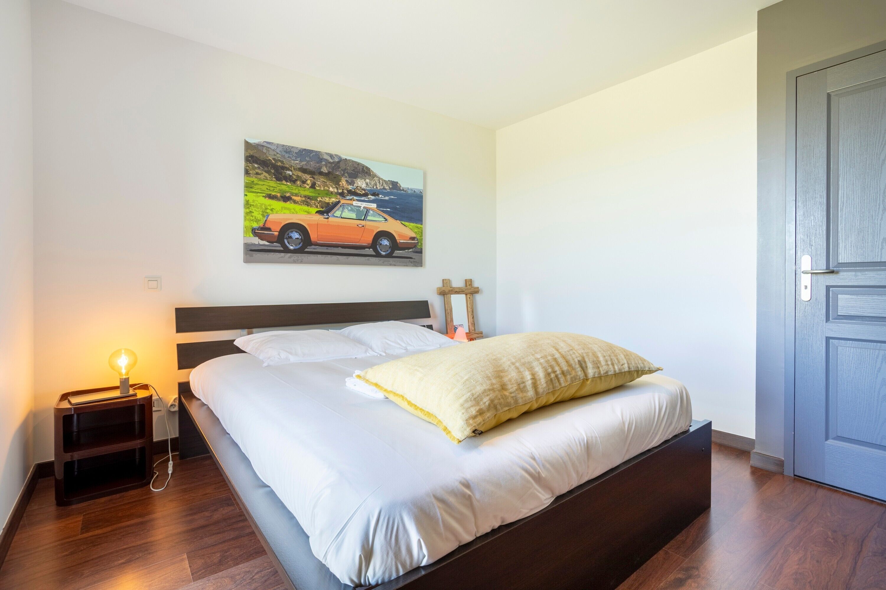 A bedroom with a bed, a nightstand with a lamp, a painting of a car, and a door.