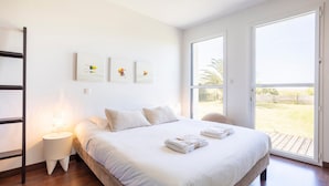 A bedroom with a large bed, a bedside table, a window, and artwork on the wall. - Maison Horizon – 3 Bedrooms/6 Guests – Sea View – 100m from Beach (Guilvinec)