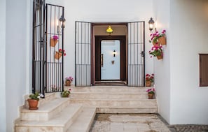 Una escalera que lleva a una puerta, con un aparato de iluminación encima, macetas a ambos lados y un farol fijado a la pared a la derecha.