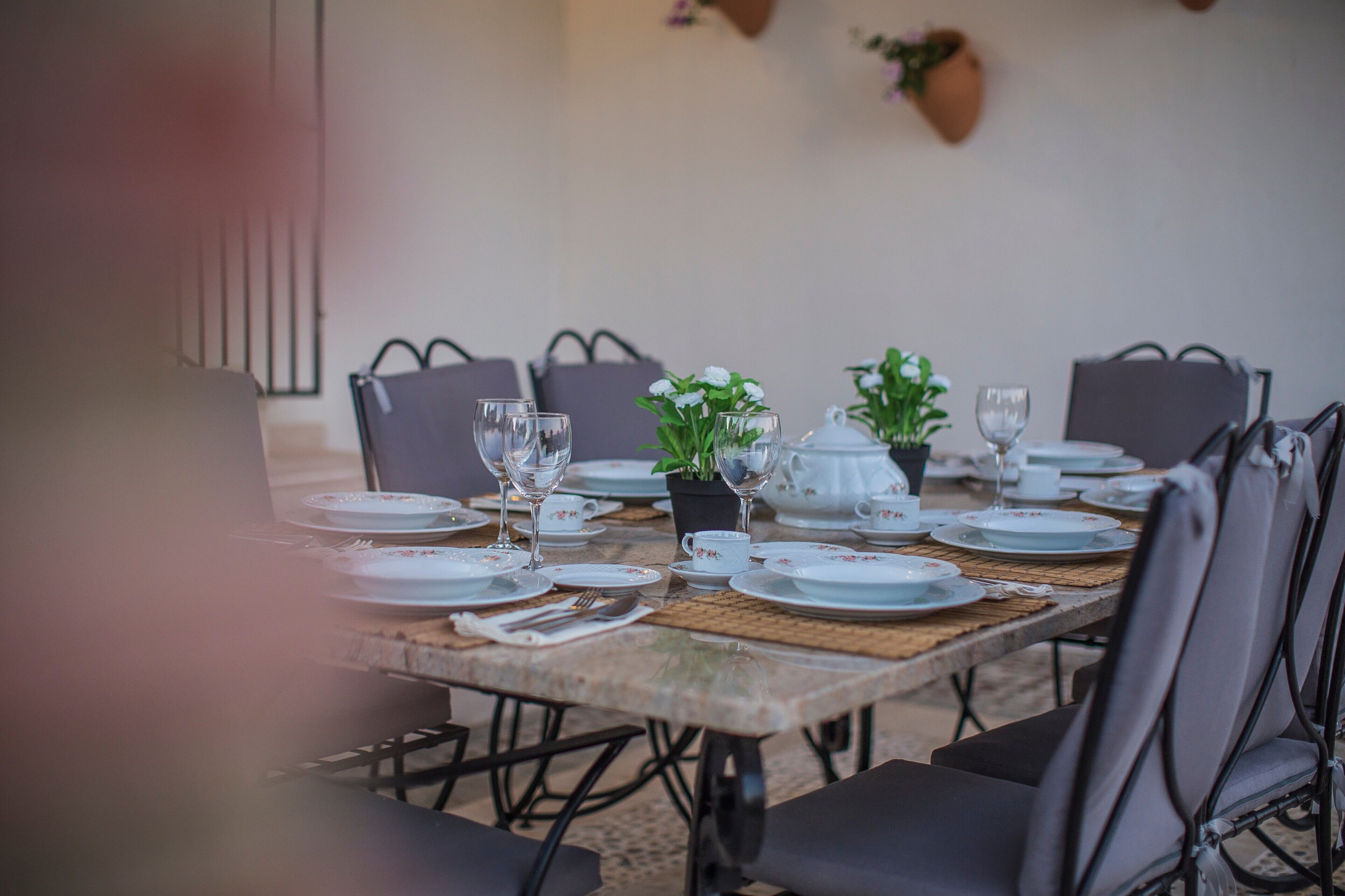 Una mesa de comedor dispuesta con platos, vasos y una tetera, rodeada de sillas.