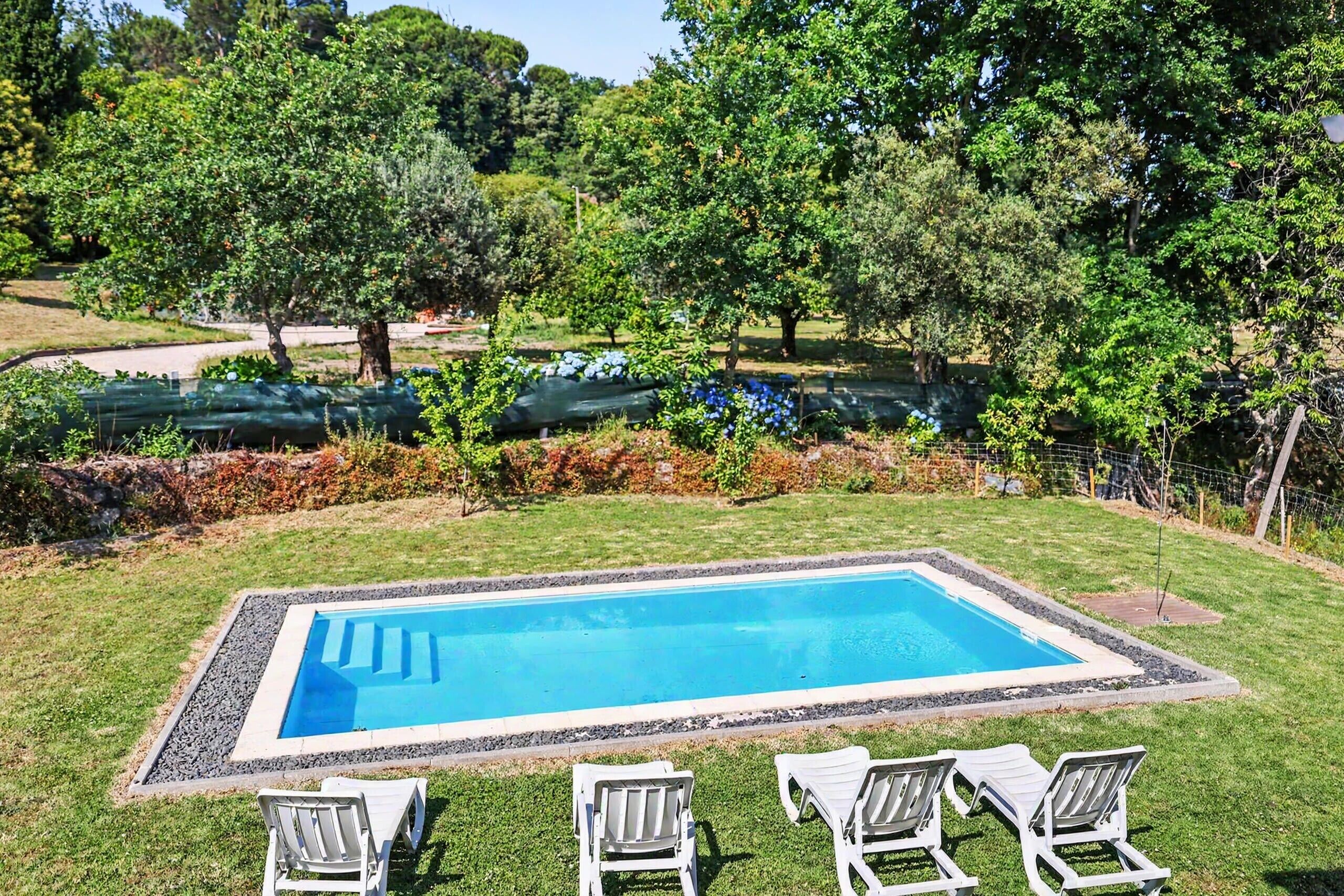 Una piscina rectangular con superficie de agua azul cristalina, rodeada por un borde de grava. Hay tres sillas de descanso blancas alineadas mirando hacia la piscina. La piscina se encuentra en un área con césped y árboles y arbustos al fondo.