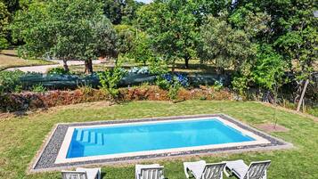 A rectangular swimming pool with a clear blue water surface, surrounded by a gravel border. There are three white lounge chairs aligned in a row facing the pool. The pool is set in a grassy area with trees and shrubs in the background.