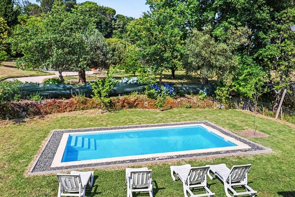 Une piscine rectangulaire à l’eau d’un bleu limpide, bordée d’un chemin de gravier. Trois chaises longues blanches sont alignées en rangée, face à la piscine. La piscine est située dans un espace verdoyant, avec des arbres et des arbustes en arrière-plan.