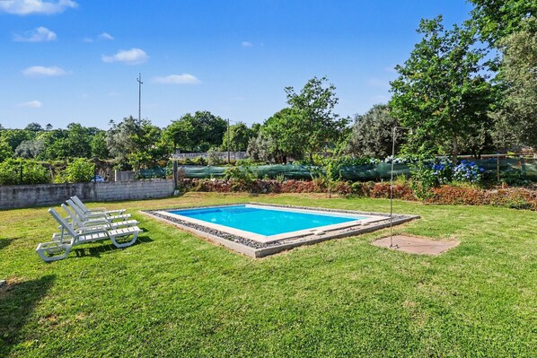 Une piscine rectangulaire entourée d’une terrasse, avec une pelouse verdoyante et une rangée de chaises longues blanches.