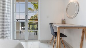 Een kamer met een schuifglasdeur die uitkomt op een terras, een klein bureau met stoel, en een ronde spiegel aan de muur.