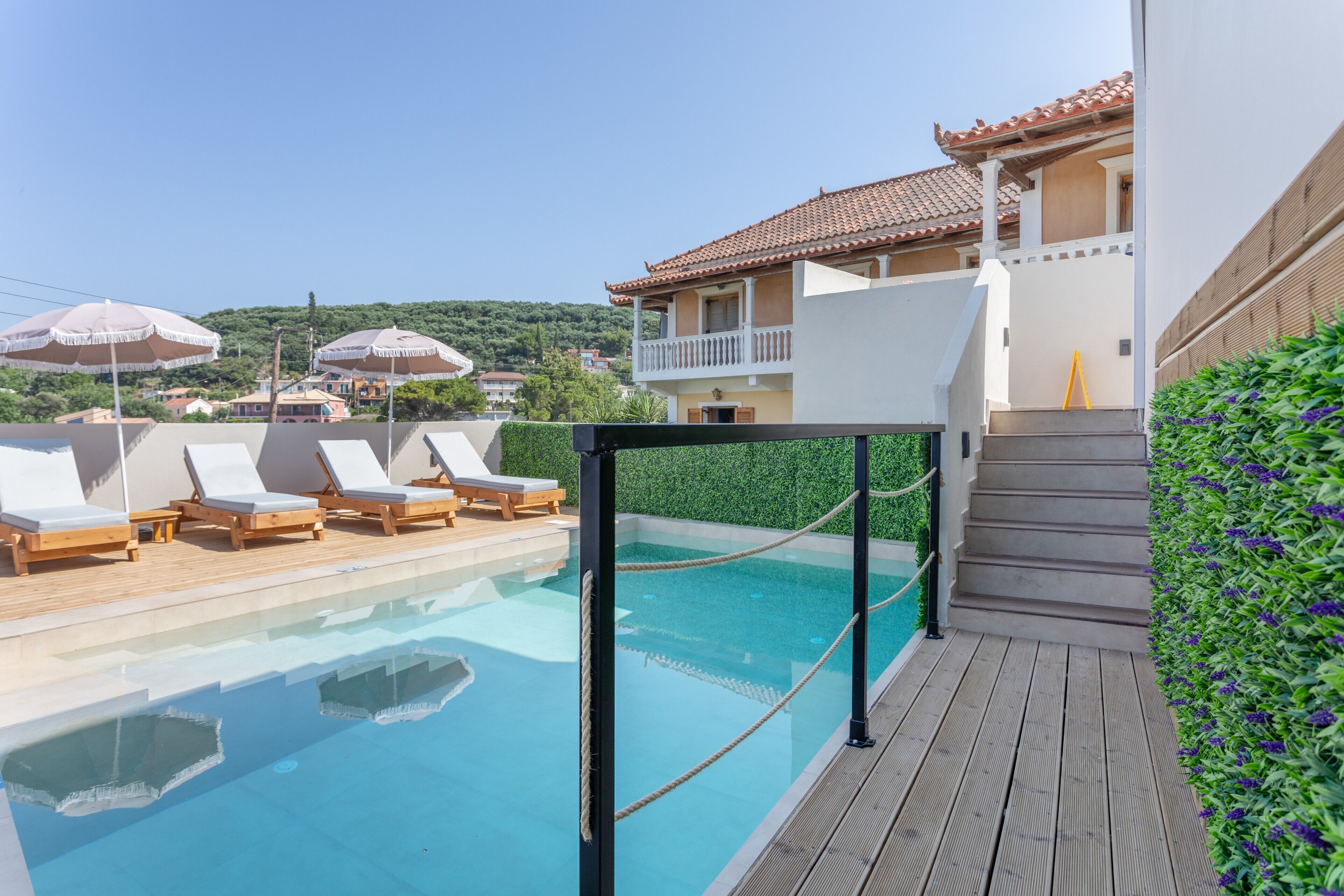 Un área de piscina con sillones de descanso y sombrillas, una terraza de madera y un edificio con balcón.