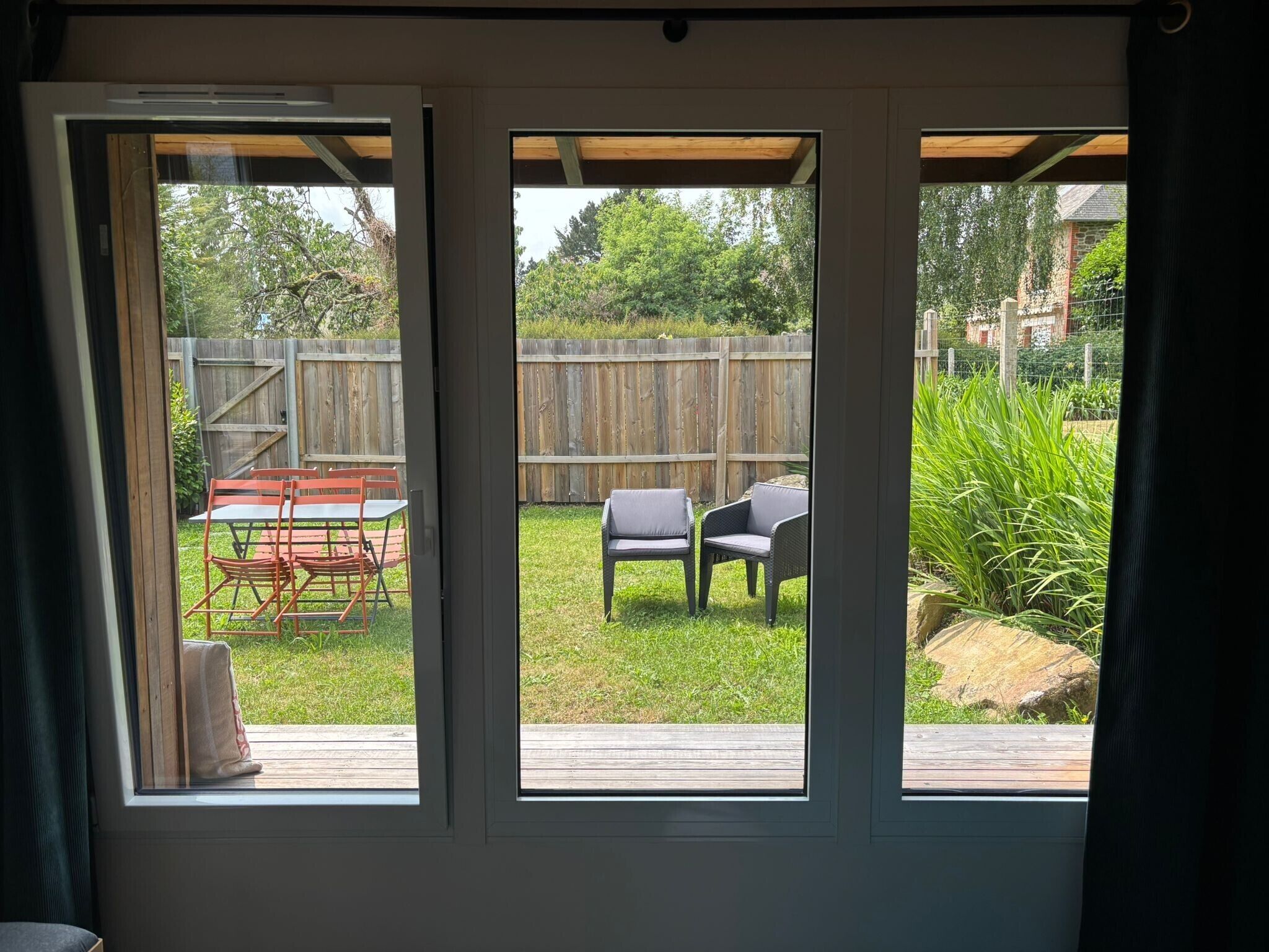 Vista a través de una ventana con una terraza de madera, muebles de exterior y una valla de madera.