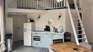A compact kitchen with a built-in oven, a refrigerator, and a sink. There is a dining table with a woven basket and a chair. A white staircase leads to an upper level. - Holiday Home “La P’tite Maison” with Private Terrace, Garden & Wi-Fi (Le Minihic-sur-Rance)
