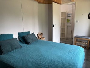 A bedroom with a bed, pillows, a bedside table, a bench, and a door leading to a bathroom. - Holiday Home 'La P’tite Maison' with Private Terrace, Garden & Wi-Fi (Le Minihic-sur-Rance)