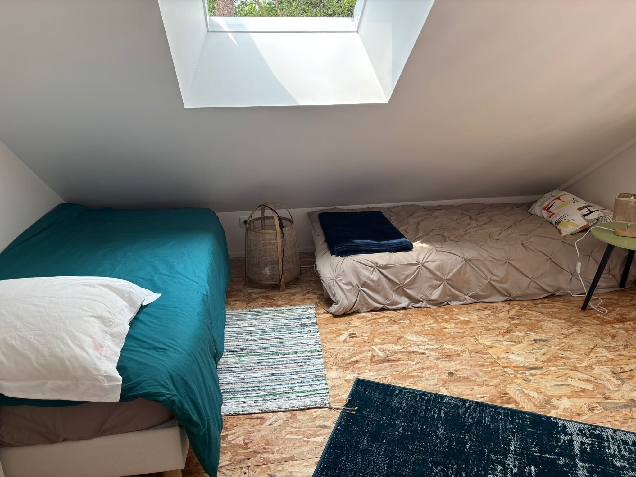 Um quarto pequeno com teto inclinado, um claraboia, uma cama com colcha azul-turquesa, um sofá com almofada branca, um cesto de vime e uma pequena mesa com abajur.