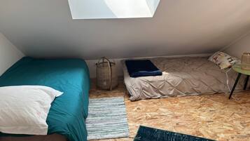 Um quarto pequeno com teto inclinado, um claraboia, uma cama com colcha azul-turquesa, um sofá com almofada branca, um cesto de vime e uma pequena mesa com abajur.