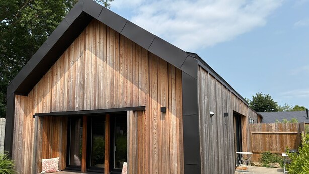 A modern wooden house with a large window, a porch with seating, and a stack of firewood outside. - Holiday Home “La P’tite Maison” with Private Terrace, Garden & Wi-Fi (Le Minihic-sur-Rance)