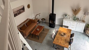 A loft-style living space with a high ceiling, a black wood-burning stove, a brown sofa, a wooden dining table, and a white cabinet. - Holiday Home “La P’tite Maison” with Private Terrace, Garden & Wi-Fi (Le Minihic-sur-Rance)