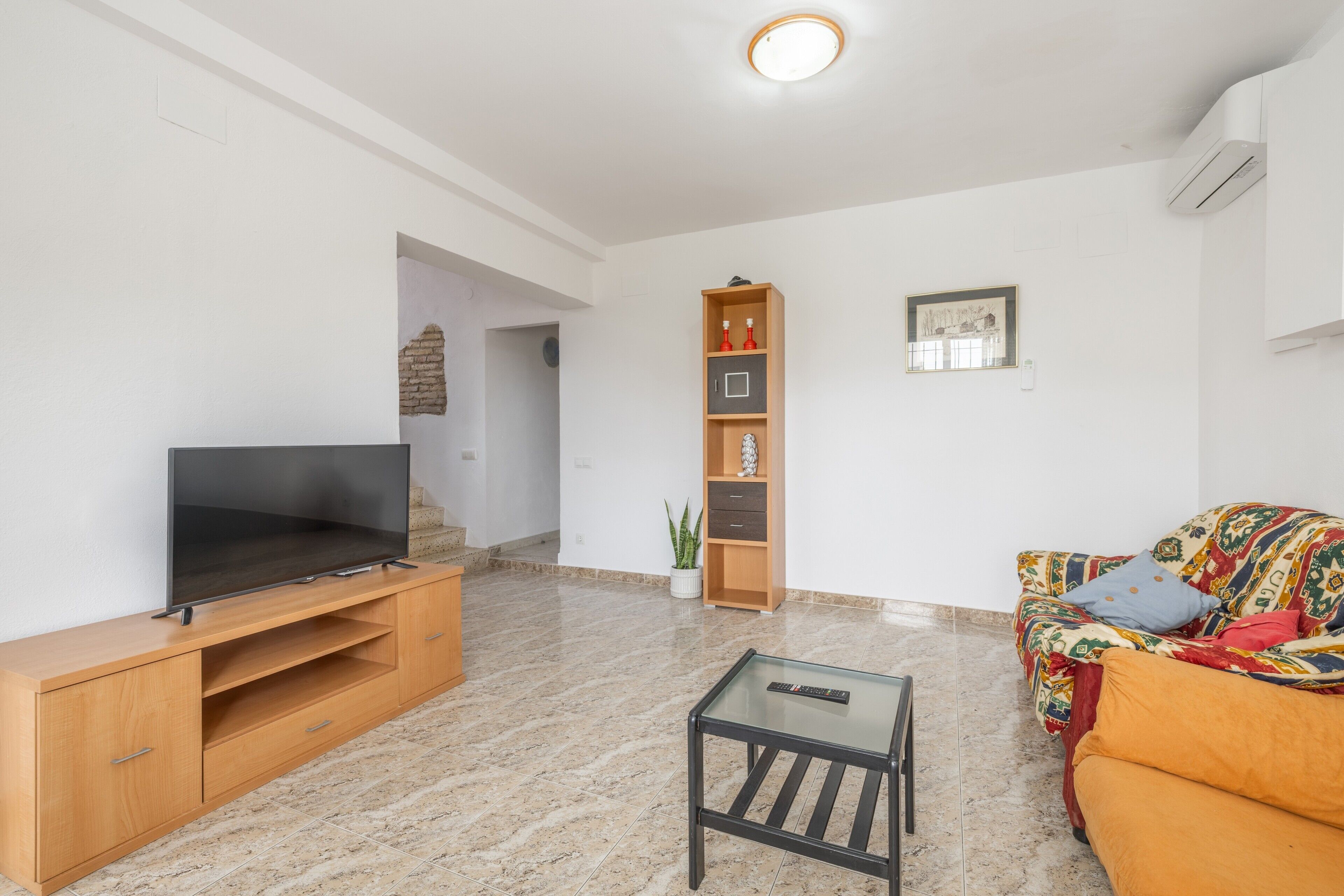 A living room with a TV on a wooden stand, a glass coffee table, a sofa with colorful cushions, and a bookshelf.