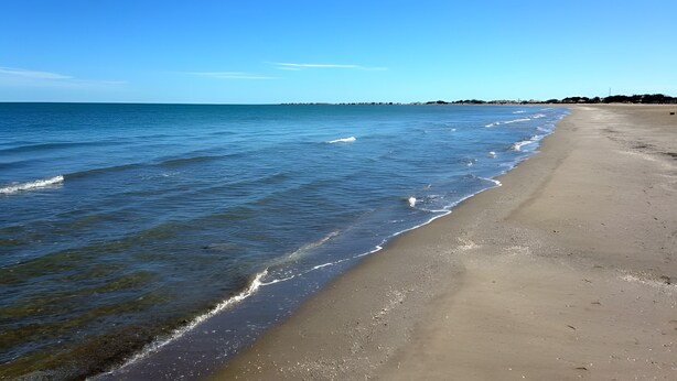 Ein Strand mit klarem, blauem Wasser, einem Sandufer und einer entfernten Küstenlinie mit Gebäuden.