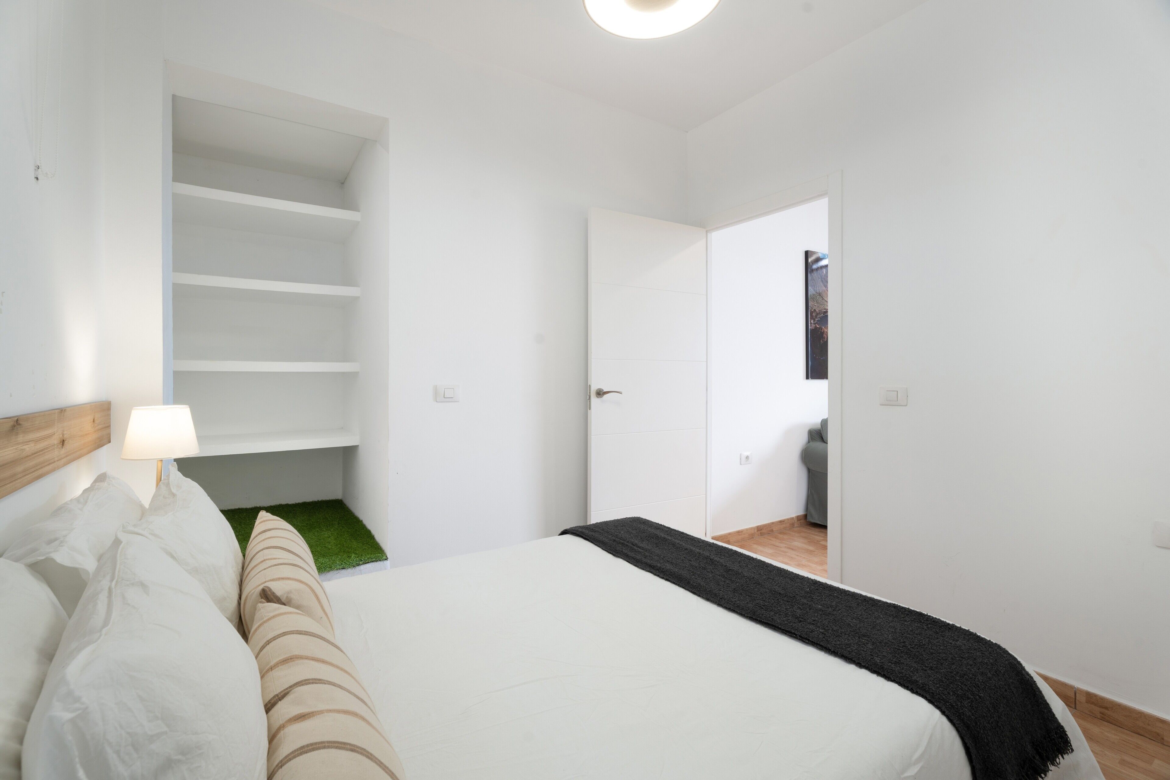 A bedroom with a bed, a bedside lamp, a green rug, a white wardrobe, and a door leading to another room.