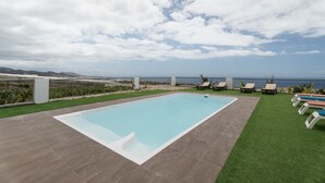A rectangular swimming pool with a white edge, surrounded by artificial grass and a tiled deck. There are lounge chairs along the pool's edge, and a view of the ocean and distant hills. - Vacation 'Beautiful Sea View and Pool Finca 1' with Shared Pool, Private Terrace and Wi-Fi (Adeje)