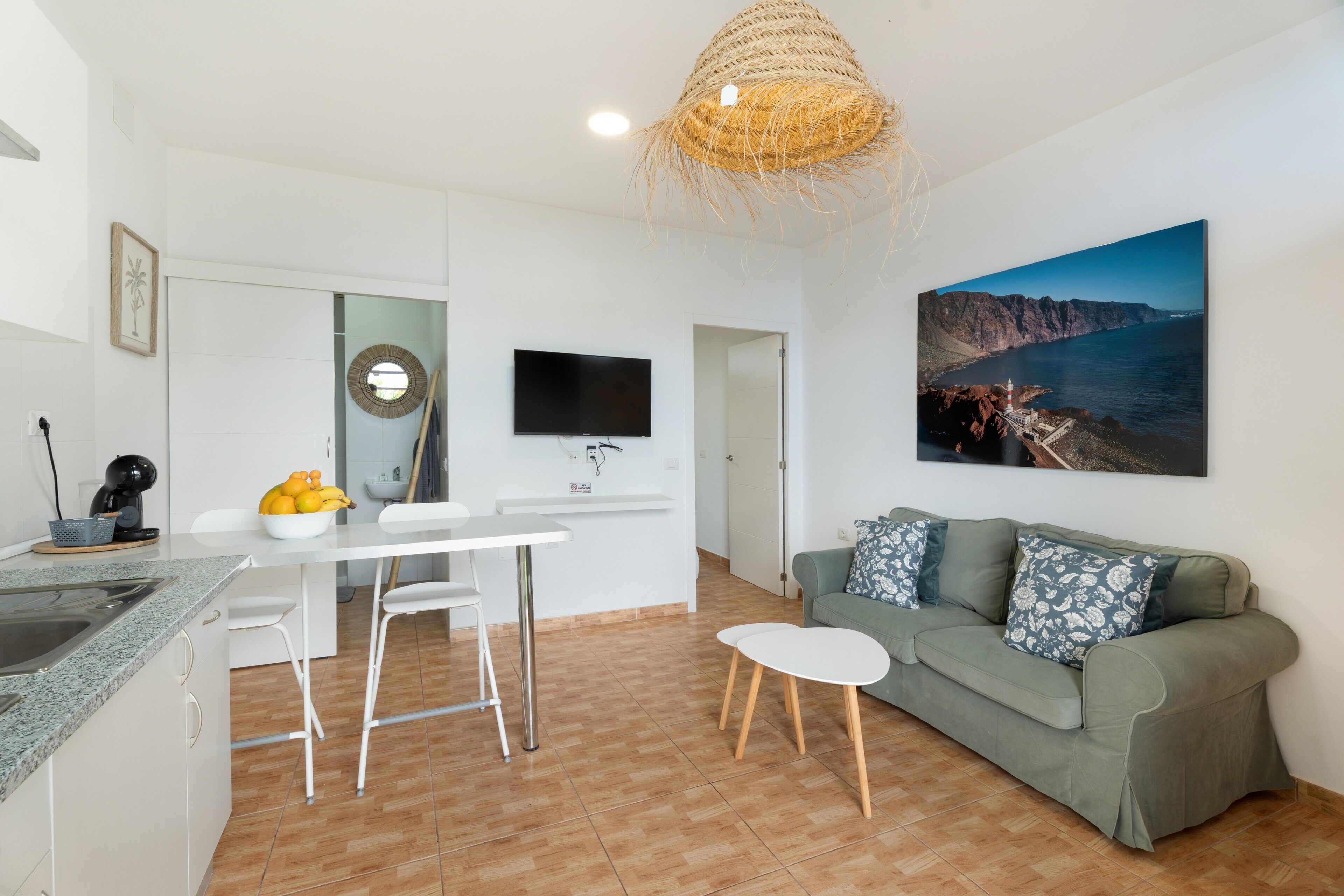 A modern kitchen with white cabinets, a dining area with a white table and chairs, a green sofa with patterned cushions, and a large painting of a coastal landscape.