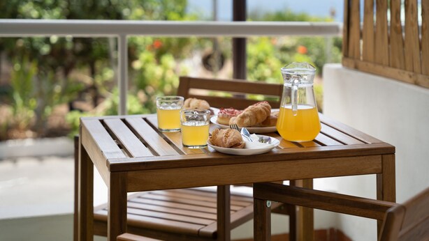 A wooden table with a glass pitcher, two glasses of orange juice, a plate with croissants, and a fork on a balcony with a view of greenery and the sea. - Vacation 'Beautiful Sea View and Pool Finca 1' with Shared Pool, Private Terrace and Wi-Fi (Adeje)