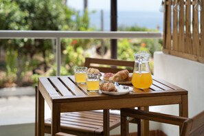 A wooden table with a glass pitcher, two glasses of orange juice, a plate with croissants, and a fork on a balcony with a view of greenery and the sea.