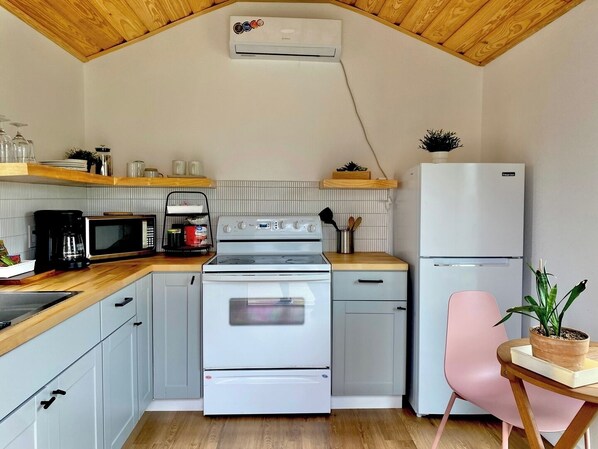 Cocina compacta con estufa, microondas y refrigerador blancos, encimeras de madera y un pequeño comedor con una silla rosa.