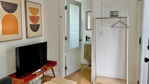A modern living room with a flat-screen TV, a wooden coffee table, and a beige ottoman. There are abstract paintings on the wall and a white door leading to a bathroom with a mirror and a towel rack. - Club Casita . Unique Stay . Easy to Airport (Austin)