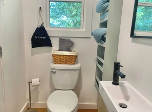 Un baño pequeño con inodoro, una cesta en la parte posterior del inodoro, un toallero con toallas, una ventana con vista a árboles y un lavamanos con grifo.