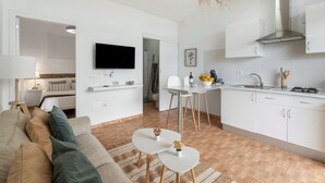 Un salon moderne et minimaliste, avec un canapé, un coin repas comprenant une table et des chaises, une cuisine dotée de meubles blancs et d’une cuisinière, et un téléviseur à écran plat fixé au mur.