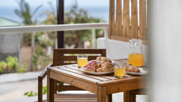 Une table en bois, dressée avec des aliments et des boissons pour le petit-déjeuner, sur un balcon donnant sur l’océan.