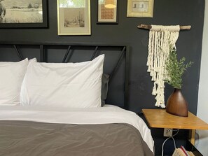 A bed with white and brown bedding, a wooden nightstand with a vase and plant, and framed artwork on the wall.