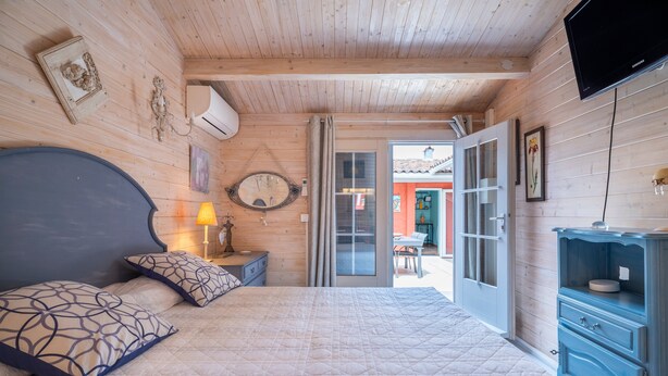 Quarto com painéis de madeira, uma cama, cômoda, televisão e porta que dá para o exterior.