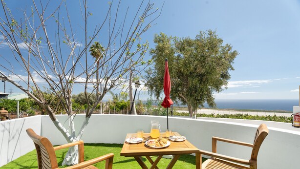 Een houten tafel met stoelen, een pitcher sap en borden met eten op een balkon met uitzicht op de oceaan.